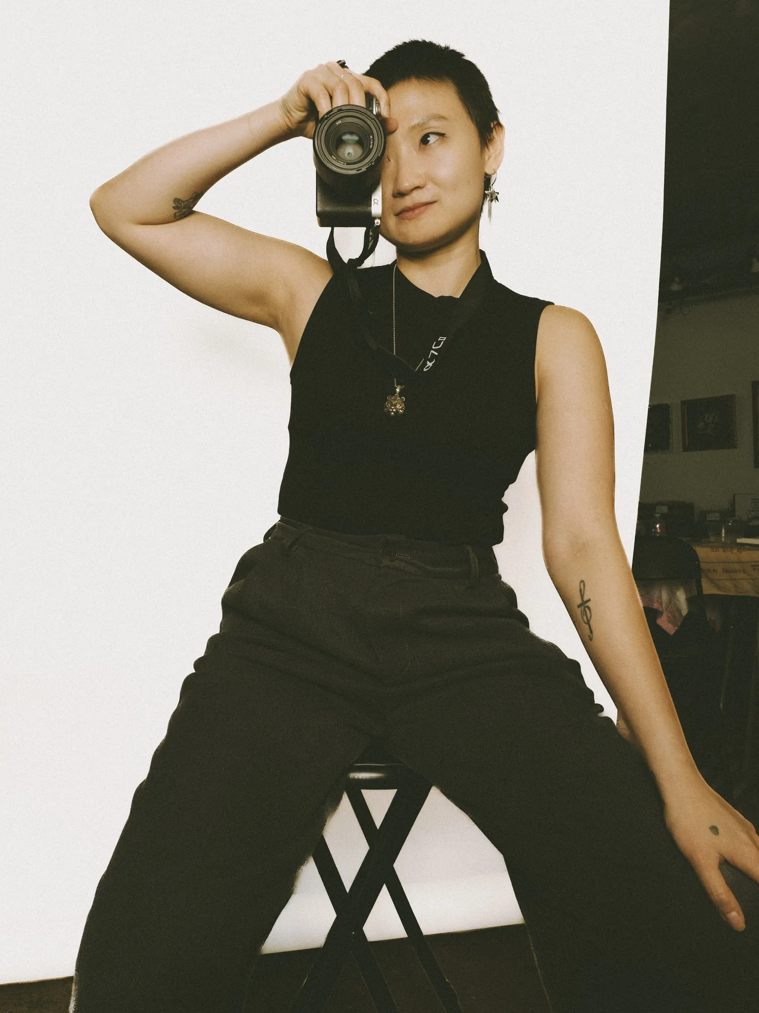 Jia seated on a stool, holding a camera up to one eye and smiling
